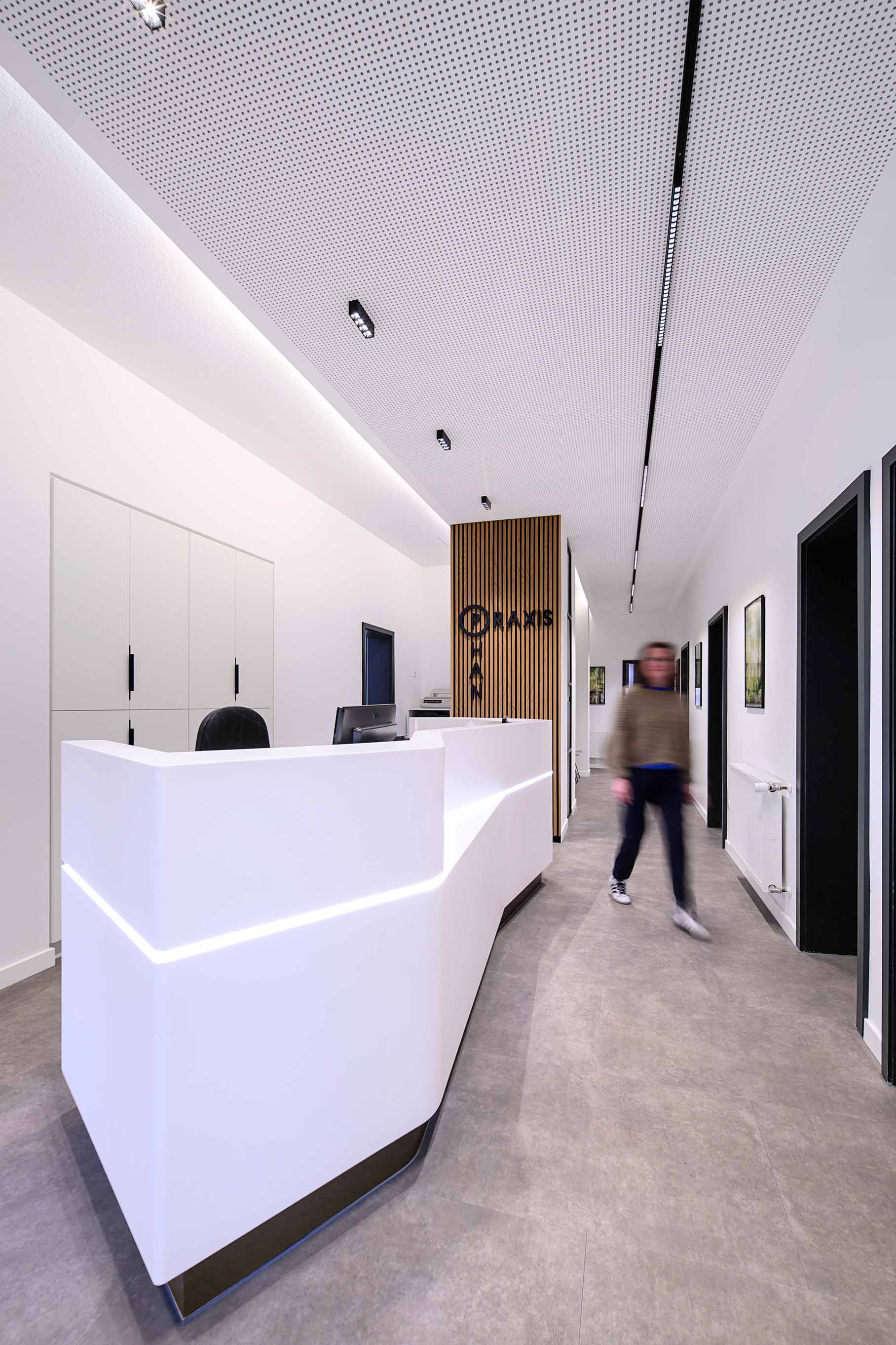 Eine Person mit unscharfem Gesichtsausdruck geht durch einen modernen weißen Empfangsbereich mit hölzernen Akzenten und heller Beleuchtung