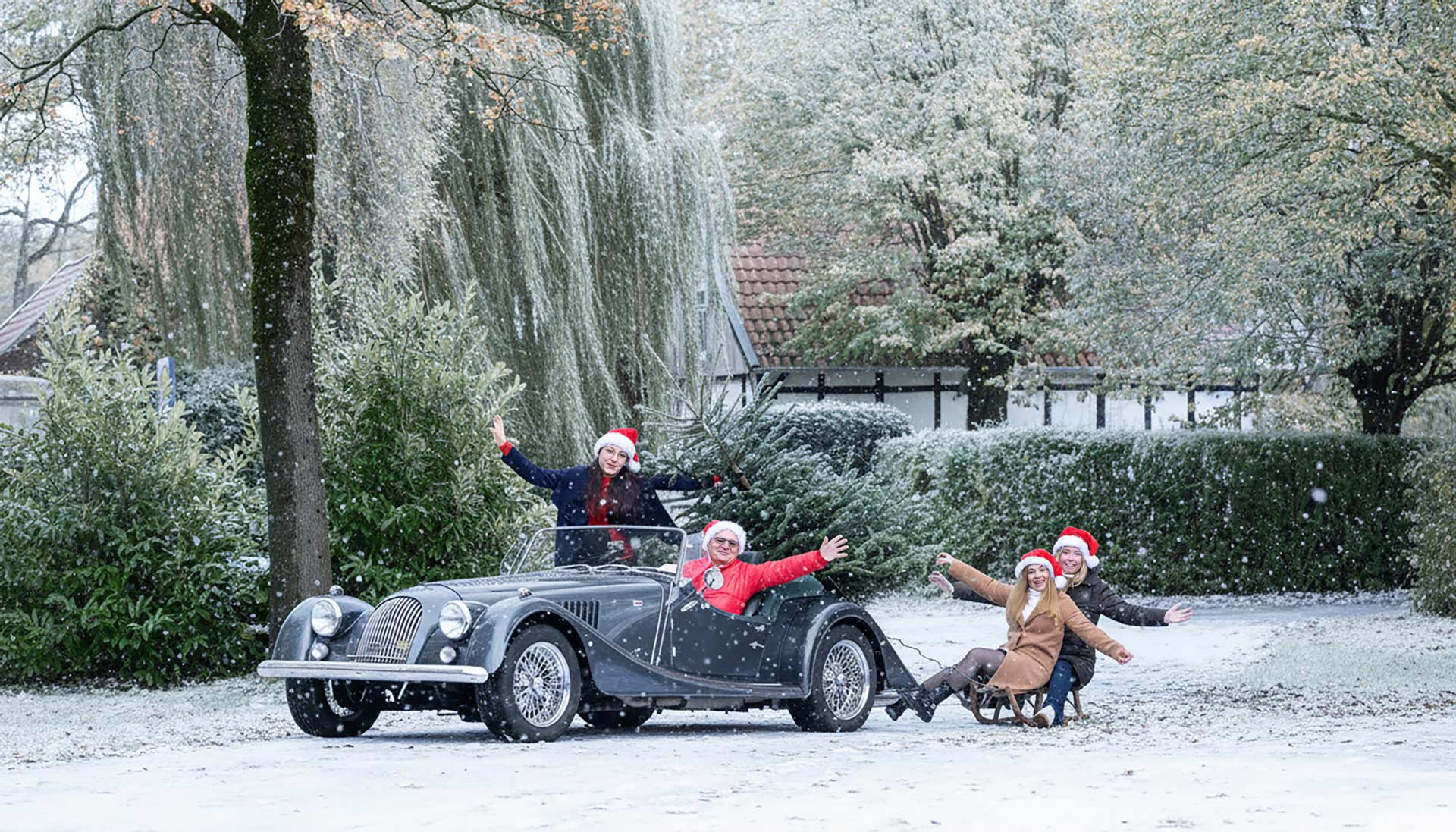 Vier fröhliche Personen mit roten Weihnachtsmützen posieren im Schnee neben einem Oldtimer