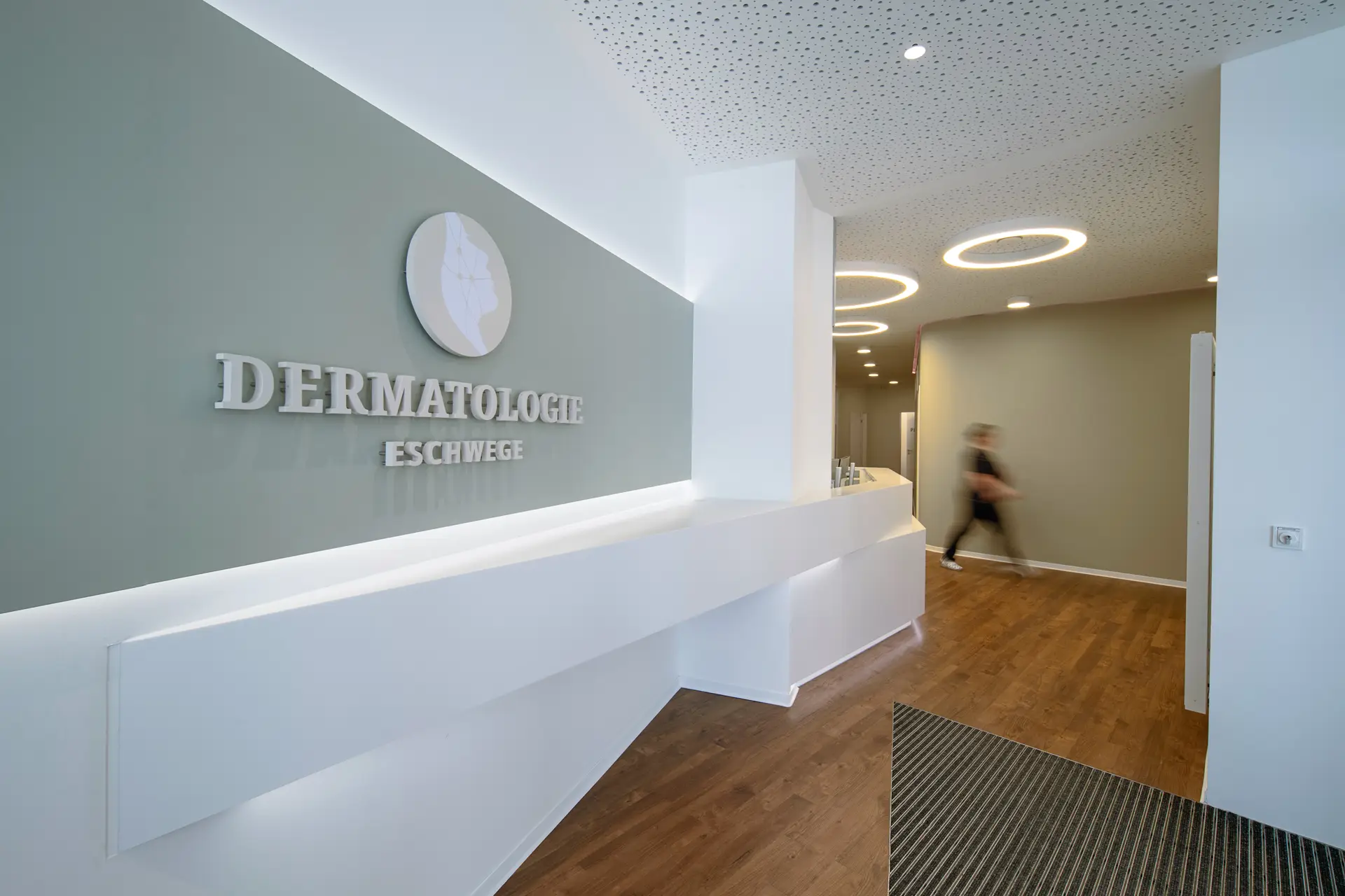 Verschwommene Person im Flur, graues Schild Dermatologie, weiße Theke, Holzboden