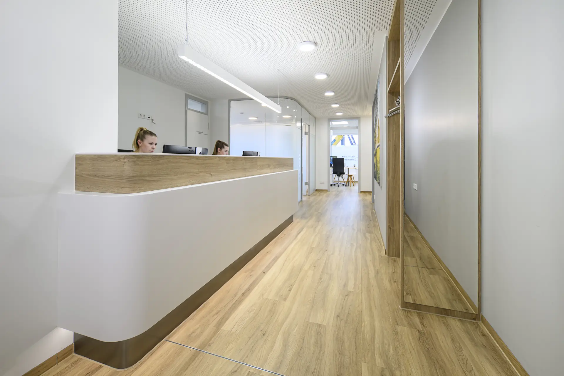 Zwei Mitarbeiterinnen mit neutralem Blick am weißen Empfang, Holzdetails, heller Flur